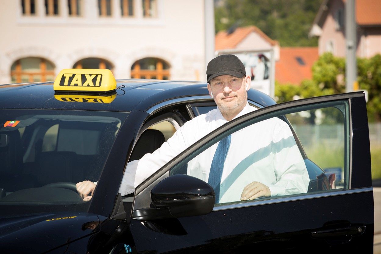 chauffeur-aba-taxi-bulle-accueil-client Chauffeur professionnel ABA Taxi à Bulle en uniforme, prêt à accueillir un client dans un véhicule noir